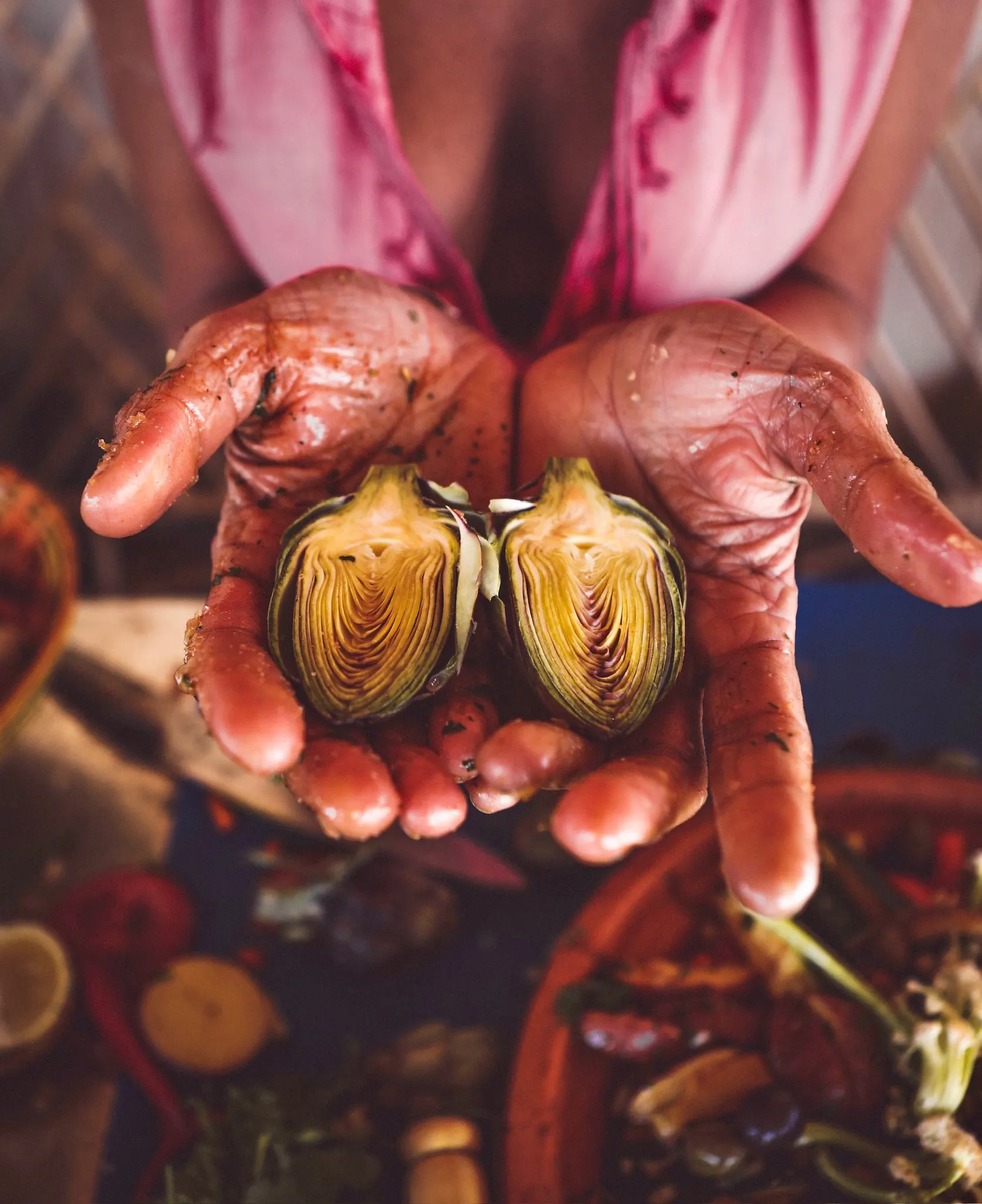 Hands glistening with spices and oil cupping a halved artichoke above a tagine