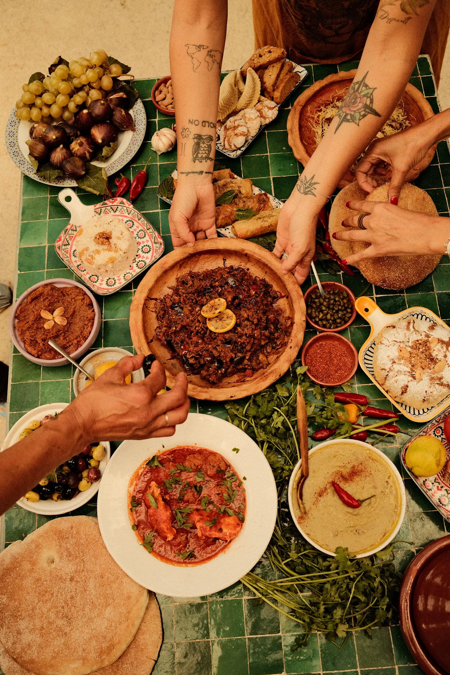 Overhead view of a Moroccan feast on a green zellige table, hands reaching for tagines, bread, figs and salads